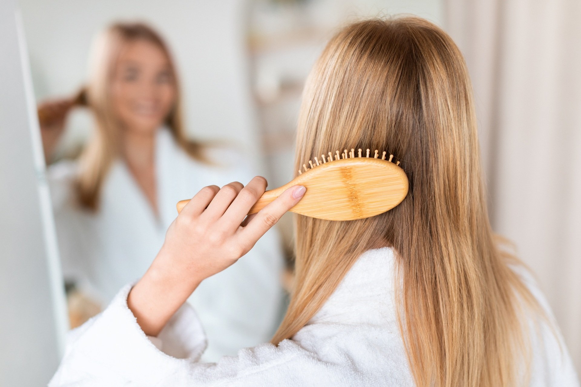 Kobieta stoi przed lustrem w łazience i rozczesuje drewnianą szczotką długie blond włosy
