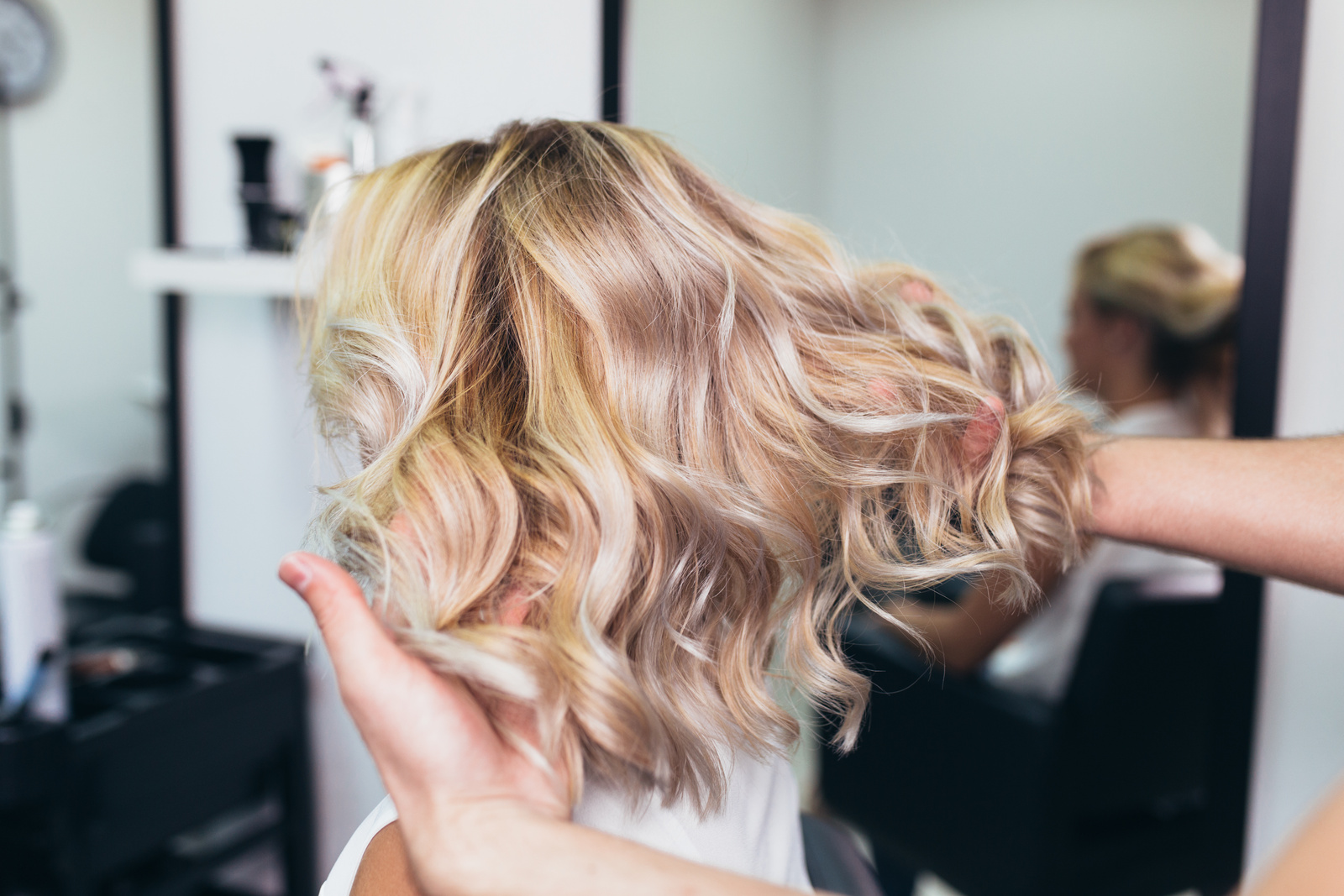 Kobieta siedzi w salonie fryzjerskim, a fryzjer układa jej blond, falowane włosy