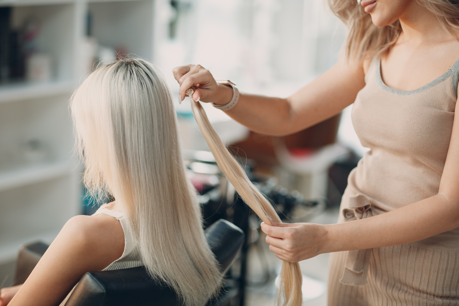Fryzjerka w salonie urody wykonuje zabieg zagęszczania i przedłużania długich, jasnych włosów klientki. Stylistka z przykłada pasma i porównuje efekt.